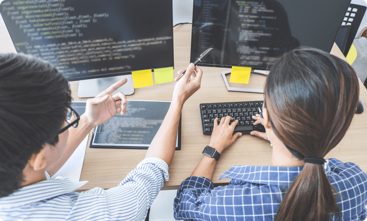 二人のプログラマーがコンピューター画面を見ながらコーディングしている。机の上には二台のモニターがあり、コードが表示されている。手前の人物はキーボードを打ち、もう一人は画面を指差している。